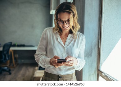 Businesswoman Using Her Smart Phone In Office. Female Entrepreneur Looking At Her Mobile Phone And Smiling. Reading Text Messages.