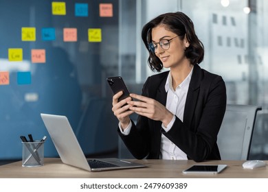 Businesswoman using smartphone at a modern office desk with laptop, demonstrating a professional working environment and multitasking capabilities. - Powered by Shutterstock