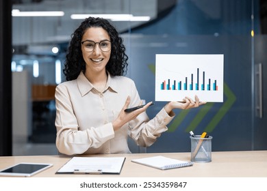 Businesswoman video call showcasing sales graph to remote team. Holding chart presenting growth. Confidently engaging digital meeting, enhancing communication. Office setting using modern technology. - Powered by Shutterstock