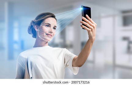Businesswoman wearing casual t-shirt is checking her personal data. Scanning her face by smart phone to unlock it. Digital connections. Concept of data protection. Blurred office on background - Shutterstock ID 2007173216