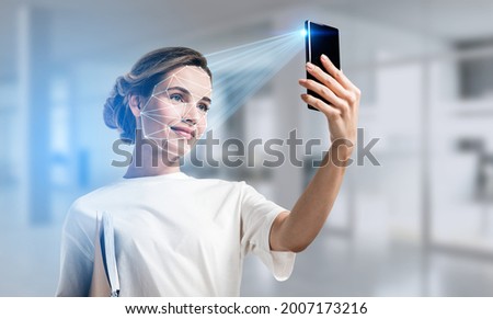 Businesswoman wearing casual t-shirt is checking her personal data. Scanning her face by smart phone to unlock it. Digital connections. Concept of data protection. Blurred office on background
