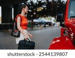 A businesswoman wearing an orange top and carrying a handbag while walking towards a red car on a city street in summer.