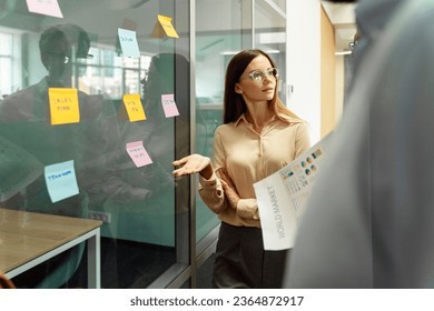 Businesswoman wearing stylish eyeglasses using sticky notes, agile methodology for productivity working together in modern office. Scrum master planning strategy, talking with business colleagues - Shutterstock ID 2364872917