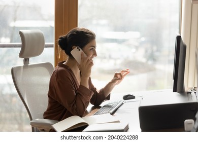 Businesswoman at work. Side shot of confident indian female manager talk on phone at workplace look on computer screen. Smiling young biracial lady expert consult client by cell sale insurance service - Powered by Shutterstock