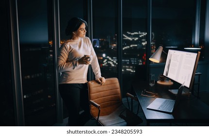 Businesswoman working late in her office, engaged in planning a social media marketing project. She stands thoughtfully in front of her desk, holding a cup of coffee and looking at her computer screen - Powered by Shutterstock