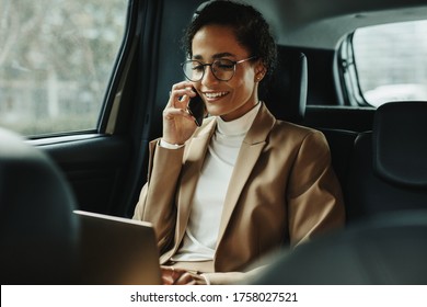 Businesswoman working on laptop and talking on phone while travelling in a taxi. Woman sitting on back seat of car using laptop and phone.
