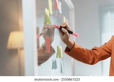 Businesswoman is writing on a transparent whiteboard with a pink marker pen in an office with post it notes stuck to it - Powered by Shutterstock