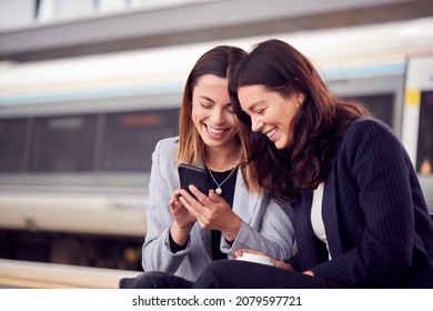 Businesswomen Commuting To Work Wait For Train On Station Platform Looking At Mobile Phone Together