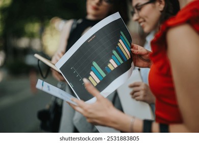 Businesswomen discussing financial data charts during an outdoor meeting. Focused on collaboration and teamwork. - Powered by Shutterstock