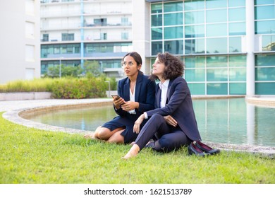 Businesswomen With Smartphone Sitting On Lawn. Serious Multiethnic Female Colleagues Resting On Green Grass And Using Cell Phone Outside Office Building. Coworkers Concept