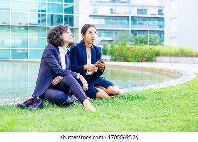 Businesswomen With Smartphone Sitting On Lawn. Serious Multiethnic Female Colleagues Resting On Green Grass And Using Cell Phone Outside Office Building. Coworkers Concept