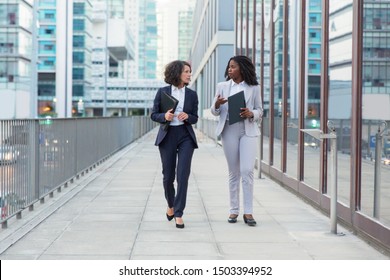 Businesswomen Walking And Talking On Street. Professional Multiethnic Female Colleagues Holding Folders, Walking And Talking Outside Office Building. Teamwork Concept