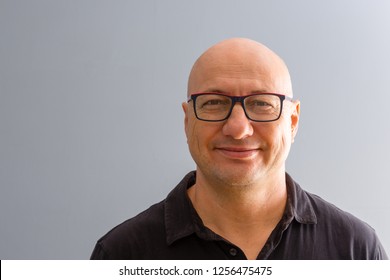 Bust Frontal Portrait Of Smiling Bold Adult Man In Glasses, Wearing Black Shirt And Standing Against Smooth Grey Background With Copy Space