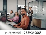 In a bustling call center, a male operator wearing glasses concentrates on his computer, diligently typing while colleagues assist customers and managers discuss strategies