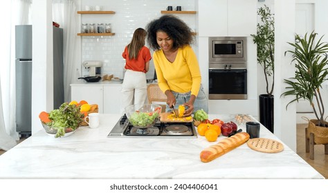 Bustling kitchen scene featuring two women engrossed in food prep. Clad in casual ware, one woman slice bell pepper on a wooden chop board, the other washing dishes. - Powered by Shutterstock