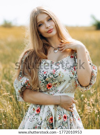 Busty pretty blonde in a beige dress with a print poses in a wheat field.