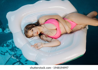 Busty sexy blonde woman in a pink swimsuit lies in the pool on a white air mattress.