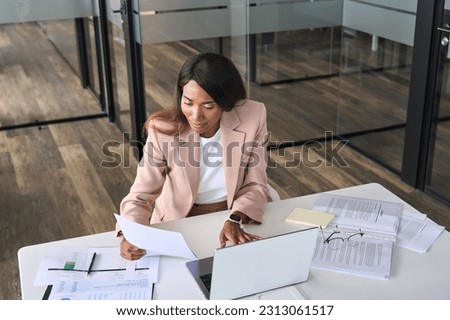 Busy African American business woman professional accountant, marketing manager, financial executive checking document report using laptop checking company papers working sitting at office desk.