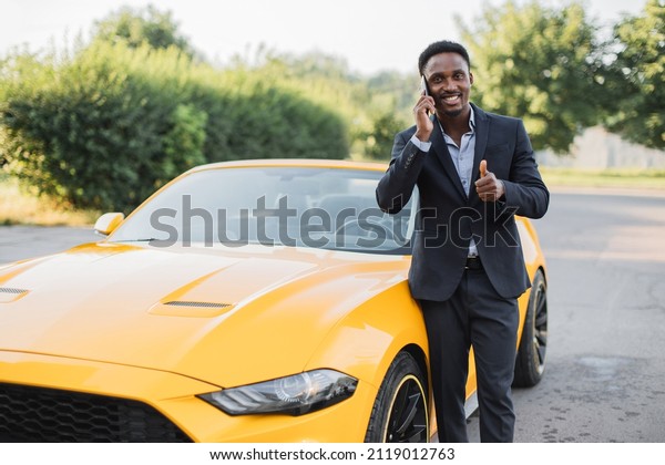 Busy african man in suit solving urgent issues on\
smartphone while standing near modern sport car. Handsome business\
man standing outdoors near his luxury yellow auto and showing thumb\
up.