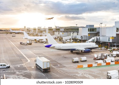 Busy airport view with airplanes and service vehicles at sunset. London airport with aircrafts at gates and taking off, trucks all around and sun setting on background. Travel and industry concepts