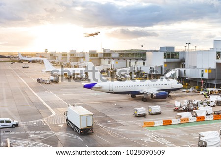 Busy airport view with airplanes and service vehicles at sunset. London airport with aircrafts at gates and taking off, trucks all around and sun setting on background. Travel and industry concepts