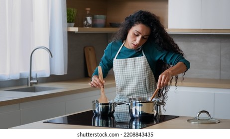 Busy Arabian Mom Attractive Curly Housewife Chef Wears Apron Cooking Breakfast At Home Kitchen Stirring Meal In Saucepans With Spoon Preparing Food For Family Talking Mobile Phone Cell Communication