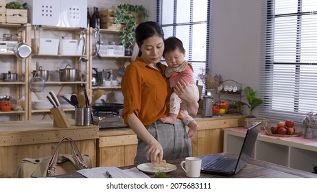 Busy Asian Career Mom Holding Baby Is Gripping The Toast From White Plate In Wooden Style Kitchen In The Morning.