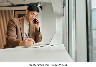 Busy Asian young business man sales manager making phone call using laptop in office. Japanese businessman talking on cellphone working consulting client discussing online digital financial data. - Powered by Shutterstock