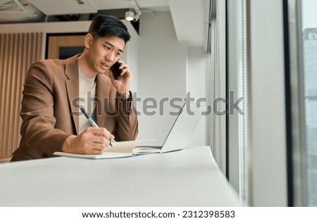 Busy Asian young business man sales manager making phone call using laptop in office. Japanese businessman talking on cellphone working consulting client discussing online digital financial data.