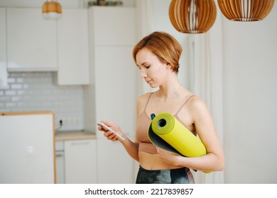 Busy Attractive Young Woman Holding A Yoga Mat, Looking At Her Phone. At Home In The Kitchen.