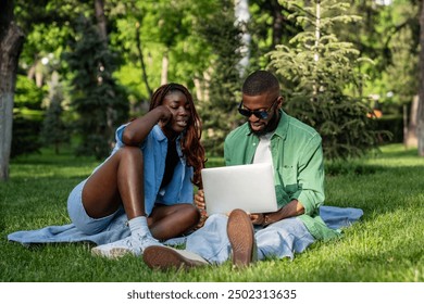 Busy black couple, male and female, engaged with laptop, viewing movies and browsing web, exchanging ideas in park on summer picnic. African students, digital nomads remote working in nature outdoors - Powered by Shutterstock