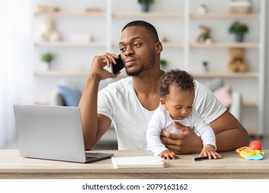 Busy Black Man Working Remotely At Home With Little Baby On Hands, African American Dad Sitting At Desk With Laptop And Talking On Cellphone While Babysitting His Infant Child, Copy Space