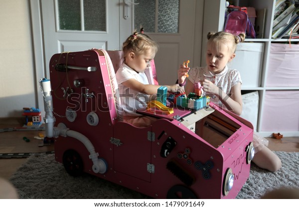 Busy\
board like a big wooden car in pink color. A little girl is playing\
with her older sister. Educational Montessori\
toys.