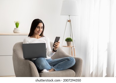 Busy business woman works with gadgets, new normal, social distance, be safe and freelance at home. Glad millennial arab lady reading message on phone in armchair, in minimalist interior with laptop