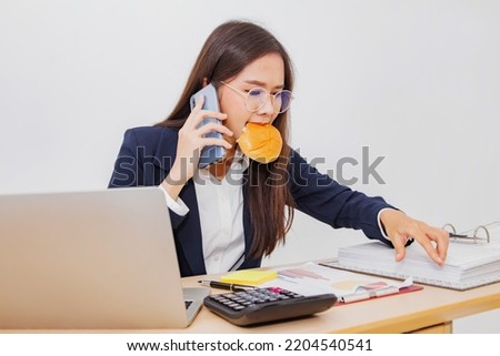 Busy businesswoman busy with work workaholic eats hamburger in her mouth while sitting at work clear documents and talk on the phone on your smartphone hear the hustle and bustle, energetic.