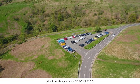 A Busy Car Park At The Bend Of A Winding Hillside Road.