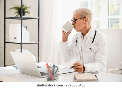 Busy caucasian elderly senior mature female doctor drinking coffee tea having breakfast while working at hospital on laptop - Powered by Shutterstock