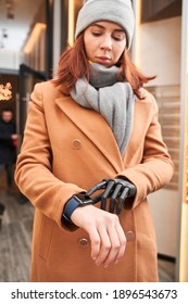 Busy caucasian female with prosthesis arm tracking time on smartwatch while standing on street near her house. Girl wearing warm clothes looking at wristwatch. Stock photo