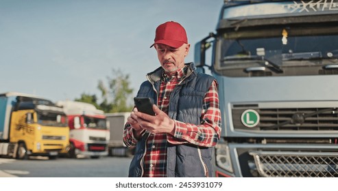 Busy Caucasian male driver chatting on his smartphone with someone about agreement on place of future delivery while standing in front of large trucks with merchandise. Transport concept.