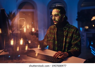 Busy Computer Specialist With Glasses In Casual Clothing Looks At His Telephone Sitting At Table With Desk Computer In Dark Office Room.