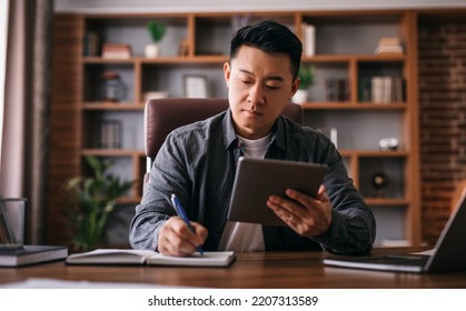 Busy Concentrated Mature Chinese Man At Table With Laptop Uses Tablet For Work In Home Office Interior. Businessman Use Application For Business Remotely, Startup And Social Distance, Ad And Offer