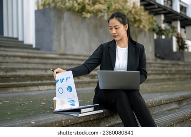 A busy, concentrated millennial Asian businesswoman in a formal business suit is checking her document and working on her laptop while sitting on stairs in front of the office building in the city. - Powered by Shutterstock