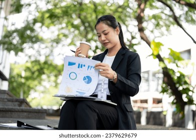 A busy, concentrated, and professional millennial Asian businesswoman is sipping coffee and reviewing business documents while sitting on the stairs in the city. - Powered by Shutterstock