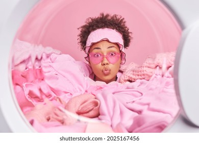 Busy curly haired housewife keeps lips folded wears trendy sunglasses loads automatic washing machine buried in heap of clothes over pink background. View from inside of washer. Home laundry service