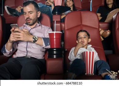 Busy Dad Looking At His Phone While Sitting In A Movie Theater With His Son