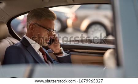Busy day. Confident and stylish mature businessman in full suit working on his laptop and talking on the phone with client while sitting in the car. Business concept