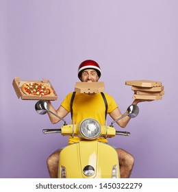 Busy deliery man drives motorbike and being overload with cardboard boxes, shows delicious pizza, has one container in mouth, wears protective helmet, isolated on purple wall with blank space above