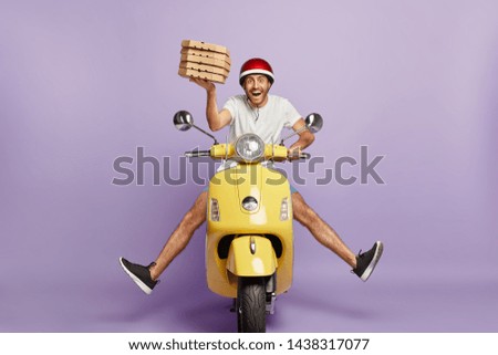Busy deliveryman being in hurry, carries cardboard boxes with pizza, delivers to customers, poses on yellow scooter, wears helmet, white t shirt and sportshoes, spreads legs, has happy expression
