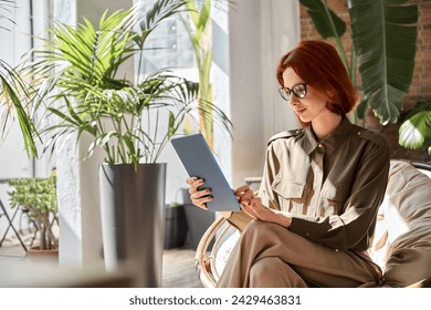 Busy elegant young professional business woman executive with red hair holding digital tablet working sitting in comfortable chair in sunny office with green plants. Candid authentic phot, copy space - Powered by Shutterstock