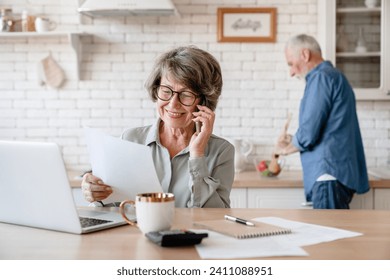 Busy european businesswoman old senior grandmother wife working, doing paperwork, dealing with documents while husband cooking dinner at home kitchen. Equality